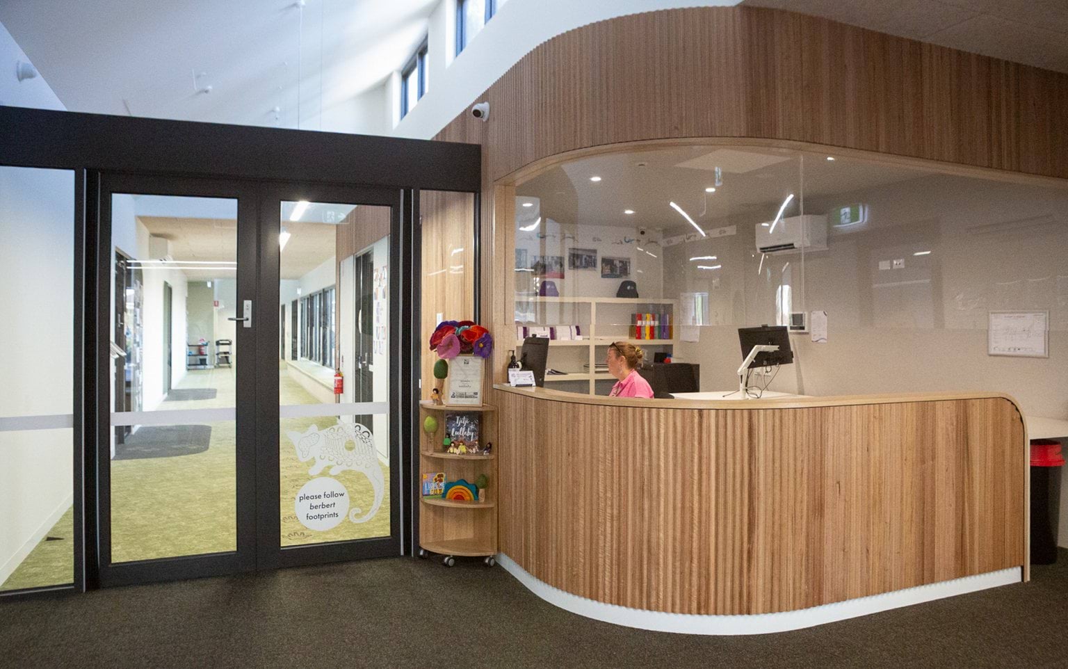Reception area at Early Learning Victoria Wimbi (Fawkner) with curved wood and glass panelling, hallway visible through glass door on left in bright, modern interior.