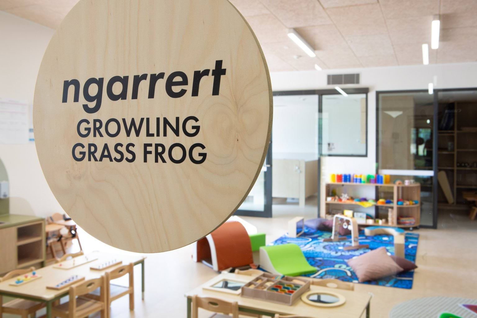 Wooden ‘Ngarrert – growling grass frog’ sign on glass door with play area visible behind at Early Learning Victoria Wimbi (Fawkner).