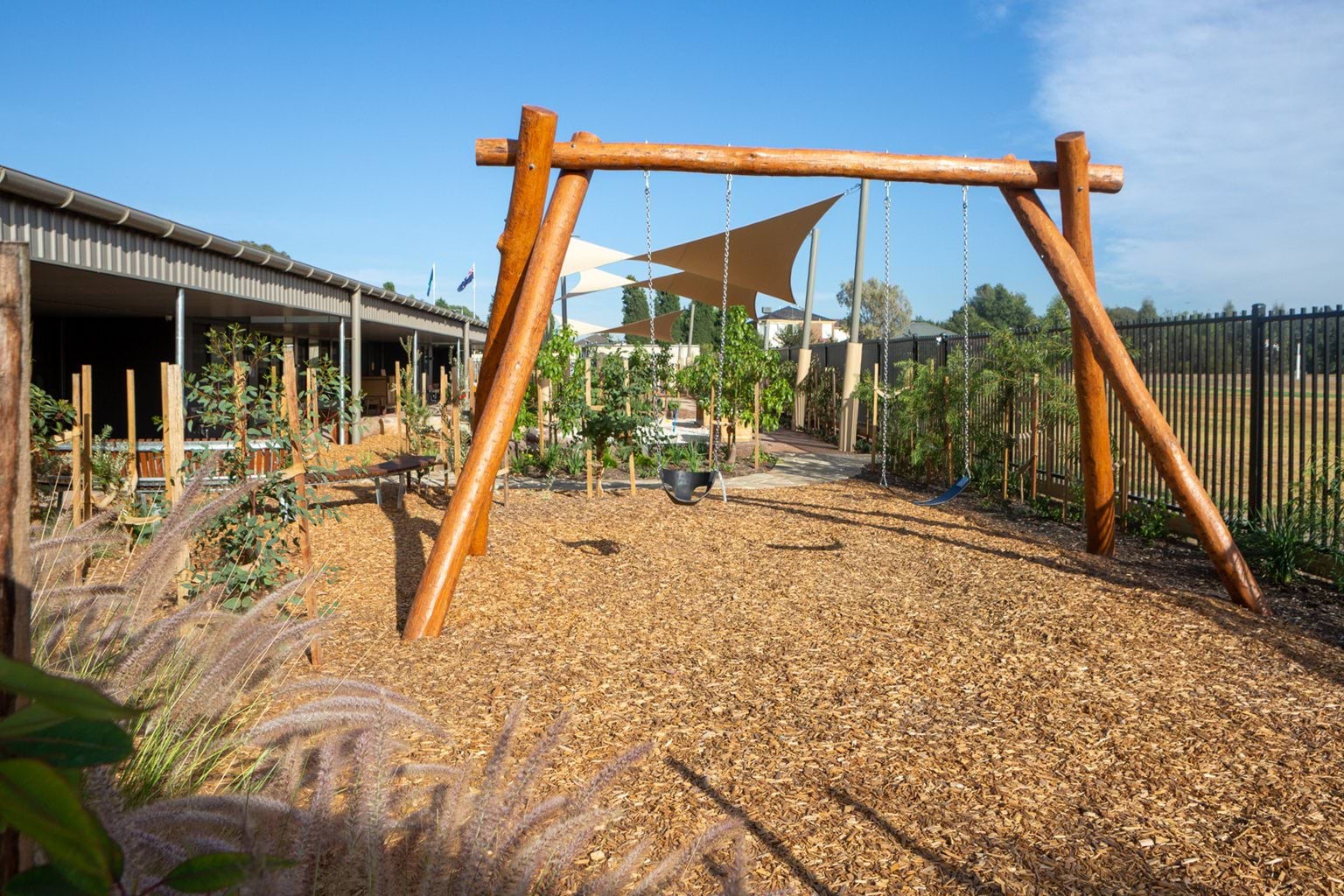 Outdoor area at Early Learning Victoria Wimbi (Fawkner) with wooden swingset, staked young plants, tanbark and shade sails under blue sky.
