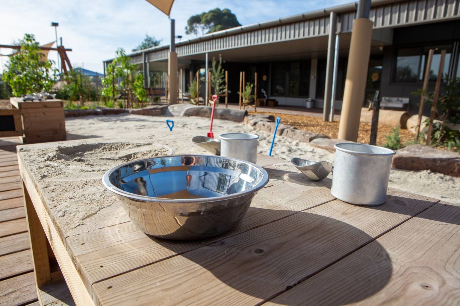 Outdoor area at Early Learning Victoria Wimbi (Fawkner) with silver bowl on timber seat and decking in foreground, sandpit with colourful shovels, trees and centre building in background.
