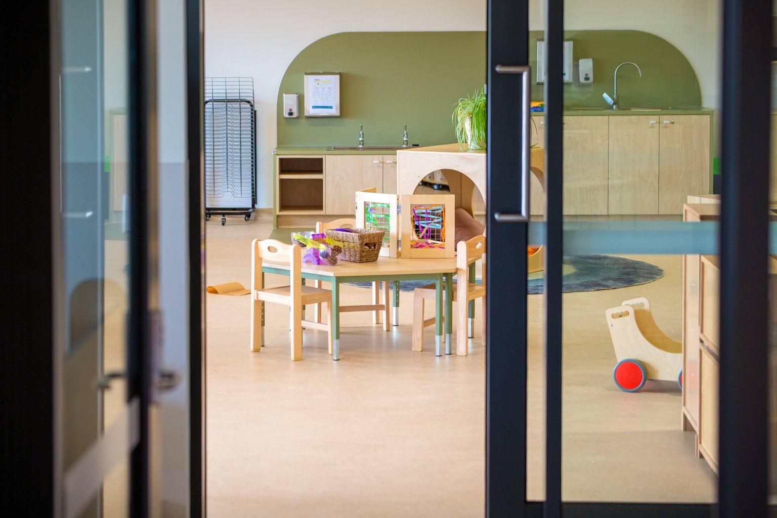 View through open glass doors into playroom at Early Learning Victoria Wimbi (Fawkner), showing timber children’s tables, chairs and toys.