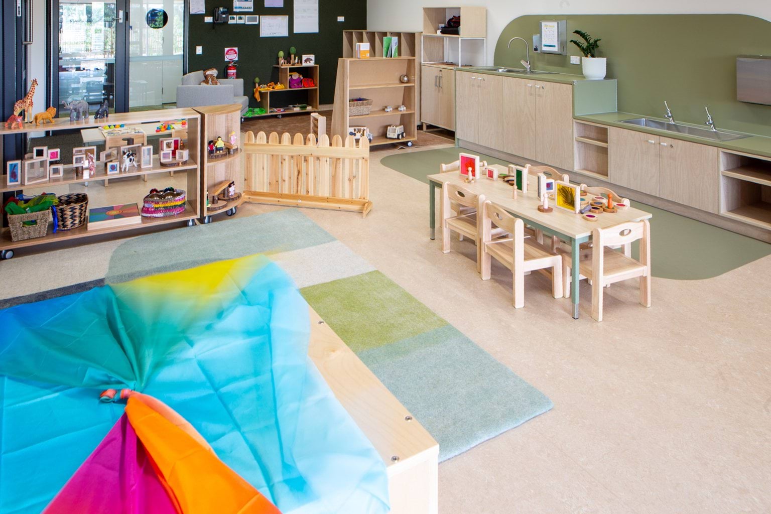 Playroom at Early Learning Victoria Wimbi (Fawkner) with colourful fabric, educational toys, mat, and wooden furniture, hallway visible in background.