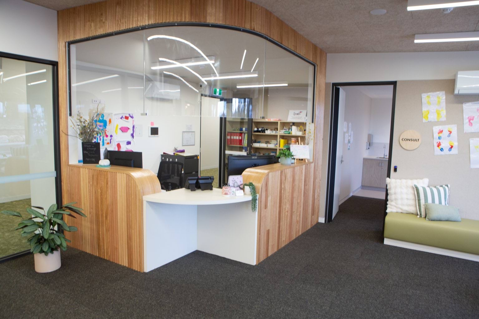 Reception area at Early Learning Victoria Nyernilang Lar (Eaglehawk North) with curved wood and glass panelling, consult room, bench with colourful cushions, children’s artwork and leafy plant in bright, modern interior.