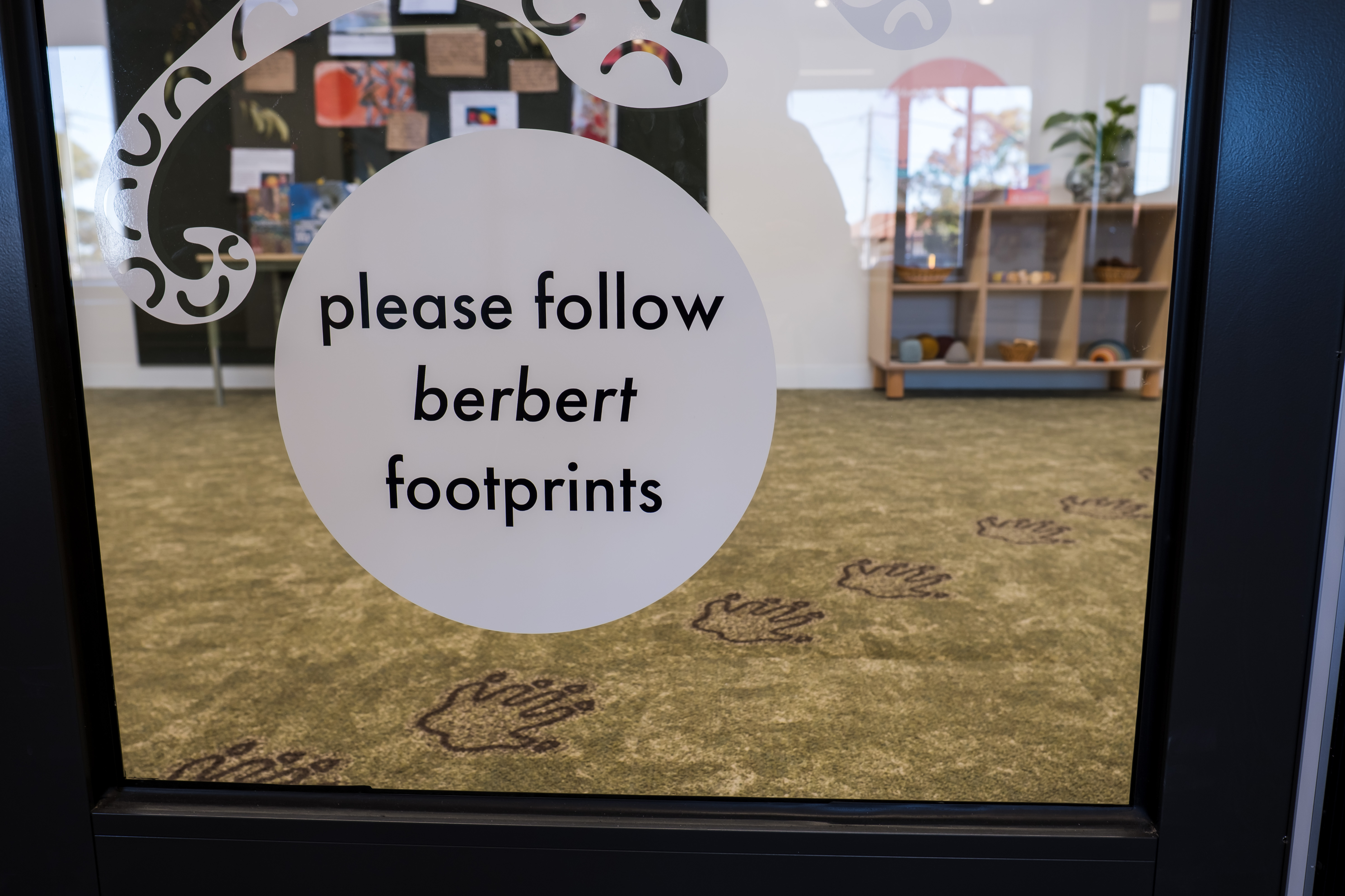 A glass door with decal sticker saying 'please follow berbert footprints', carpet behind has animal footprints leading away and to the right.