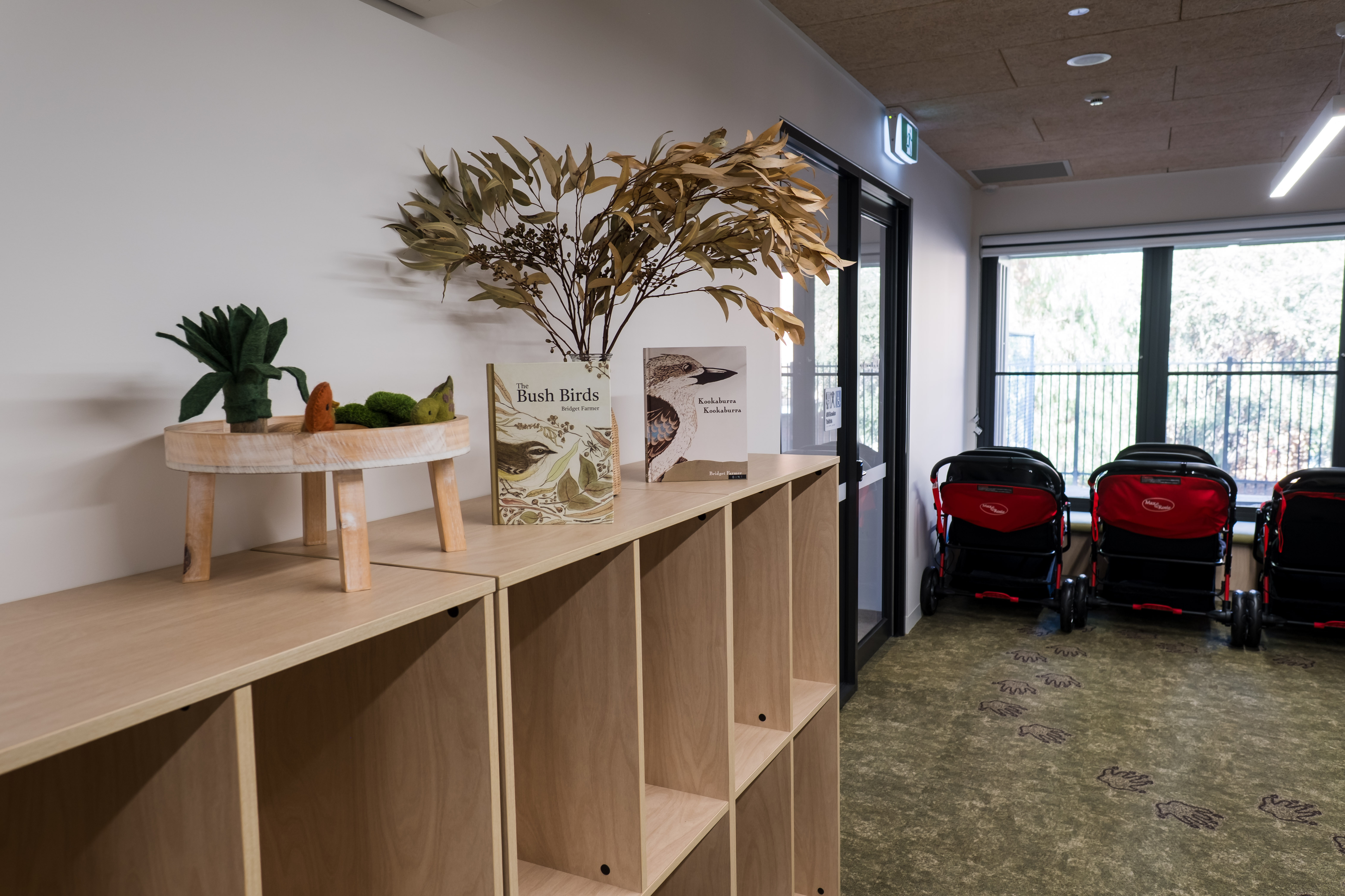 Interior close up of hallway shelves at Early Learning Victoria Muyan, showing felt toys, Bush Birds book, and a eucalyptus branch. Strollers are in the background near a window.