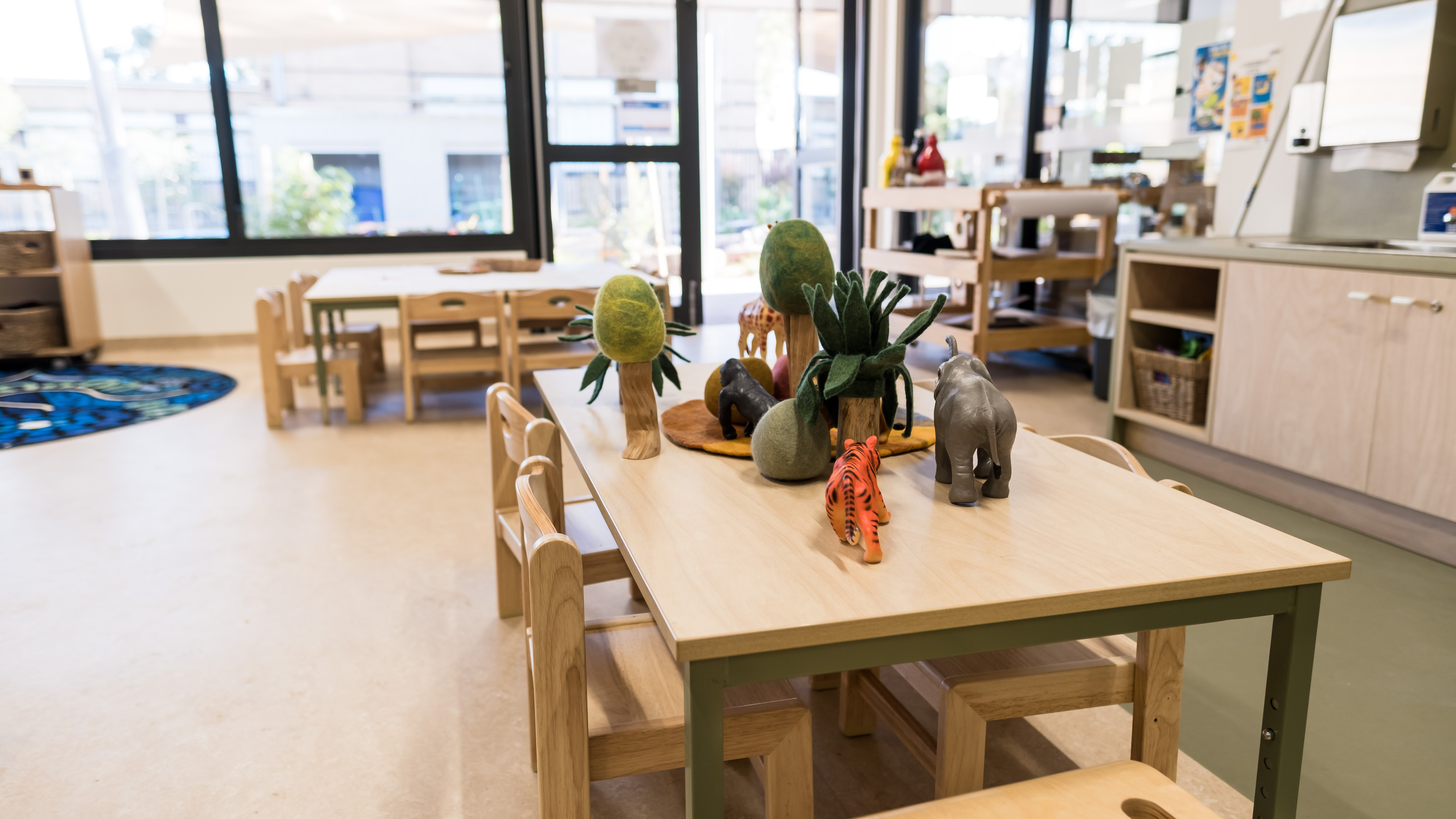 Interior view of a play room at Early Learning Victoria Muyan, with children's wooden table and chairs in foreground covered in animal toys and felt trees and rocks. Outdoor visible through windows in background.
