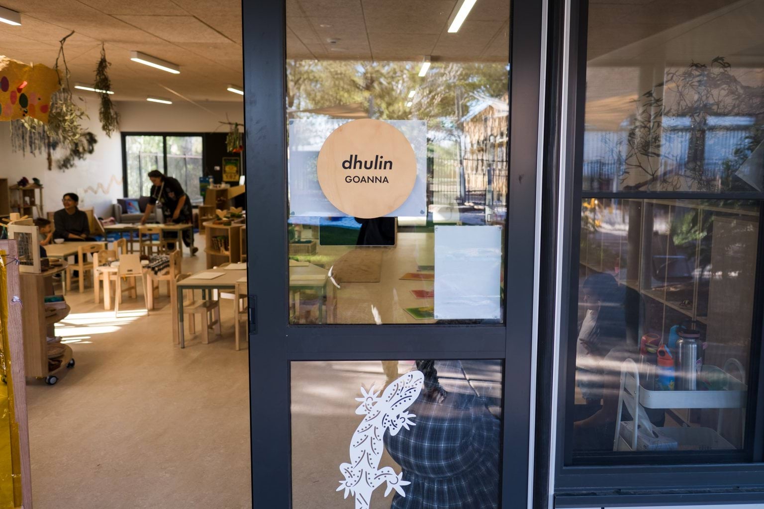 A view through the glass door of the dhulin (goanna) room at Early Learning Victoria Muyan. Light wood furniture and toys are within, and gum tree branches hand from the ceiling.